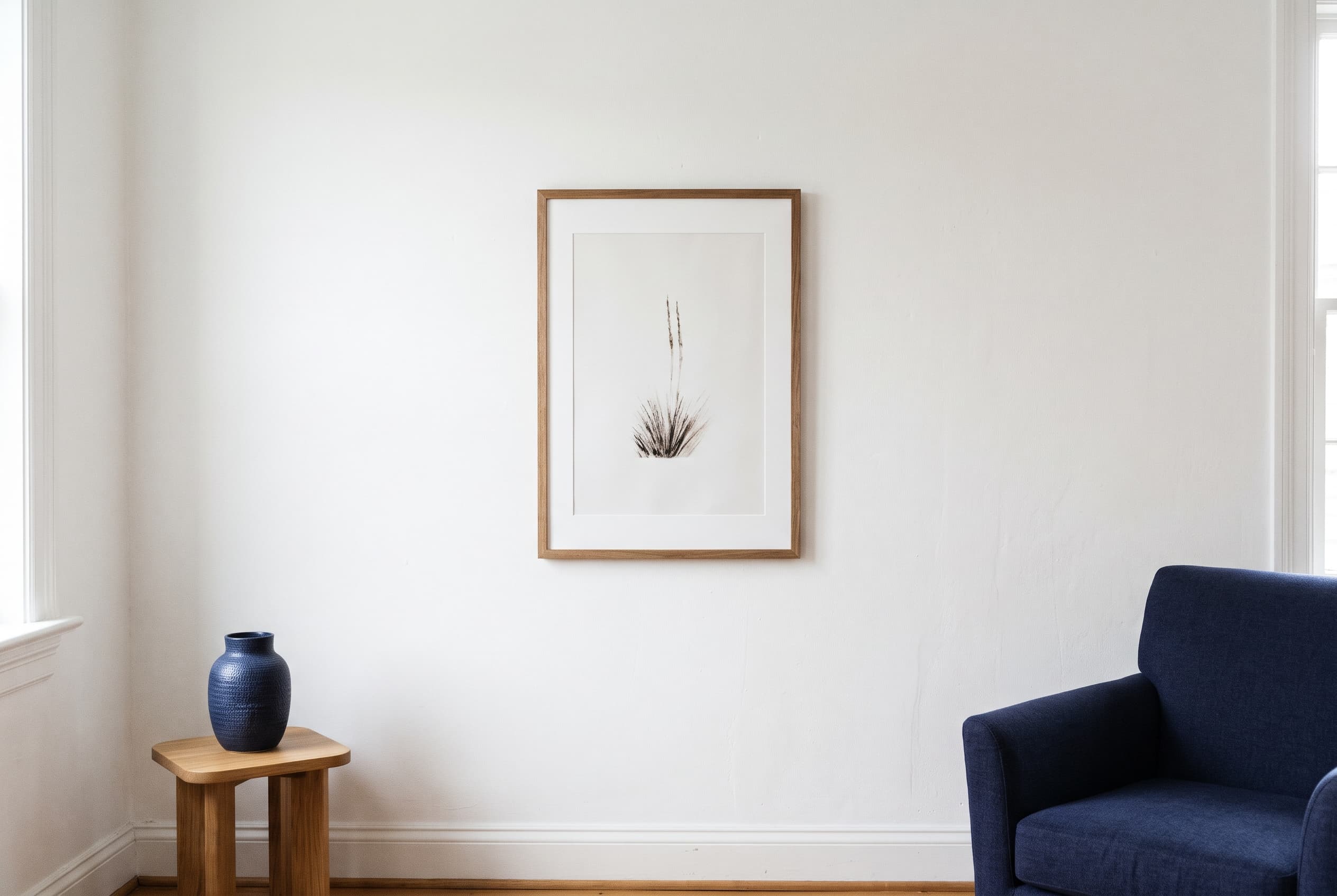 A framed print hanging level on a white wall in a quiet room, beside a navy armchair and vase.